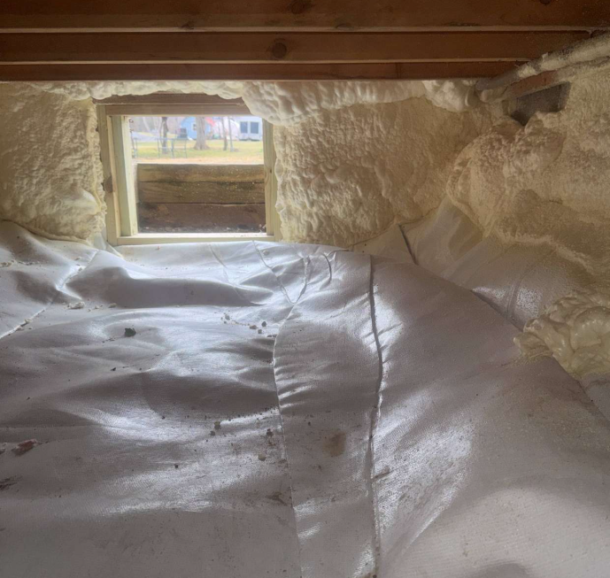 Crawl space interior lined with spray foam insulation and a white vapor barrier, with a small rectangular vent opening to the outside.