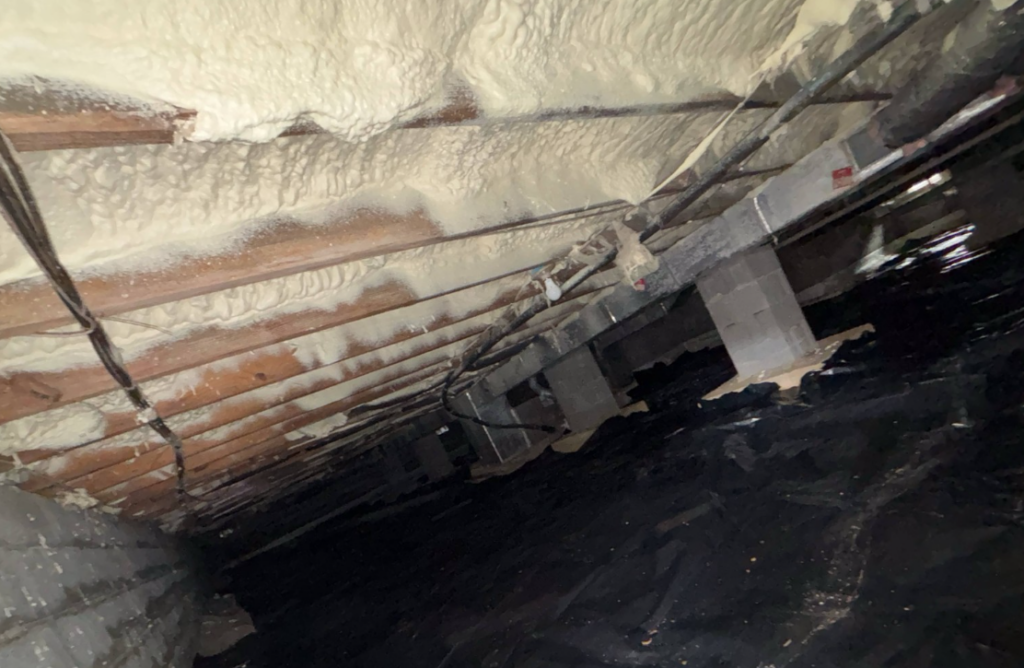 Crawl space with spray foam insulation on floor joists and plastic ground covering.